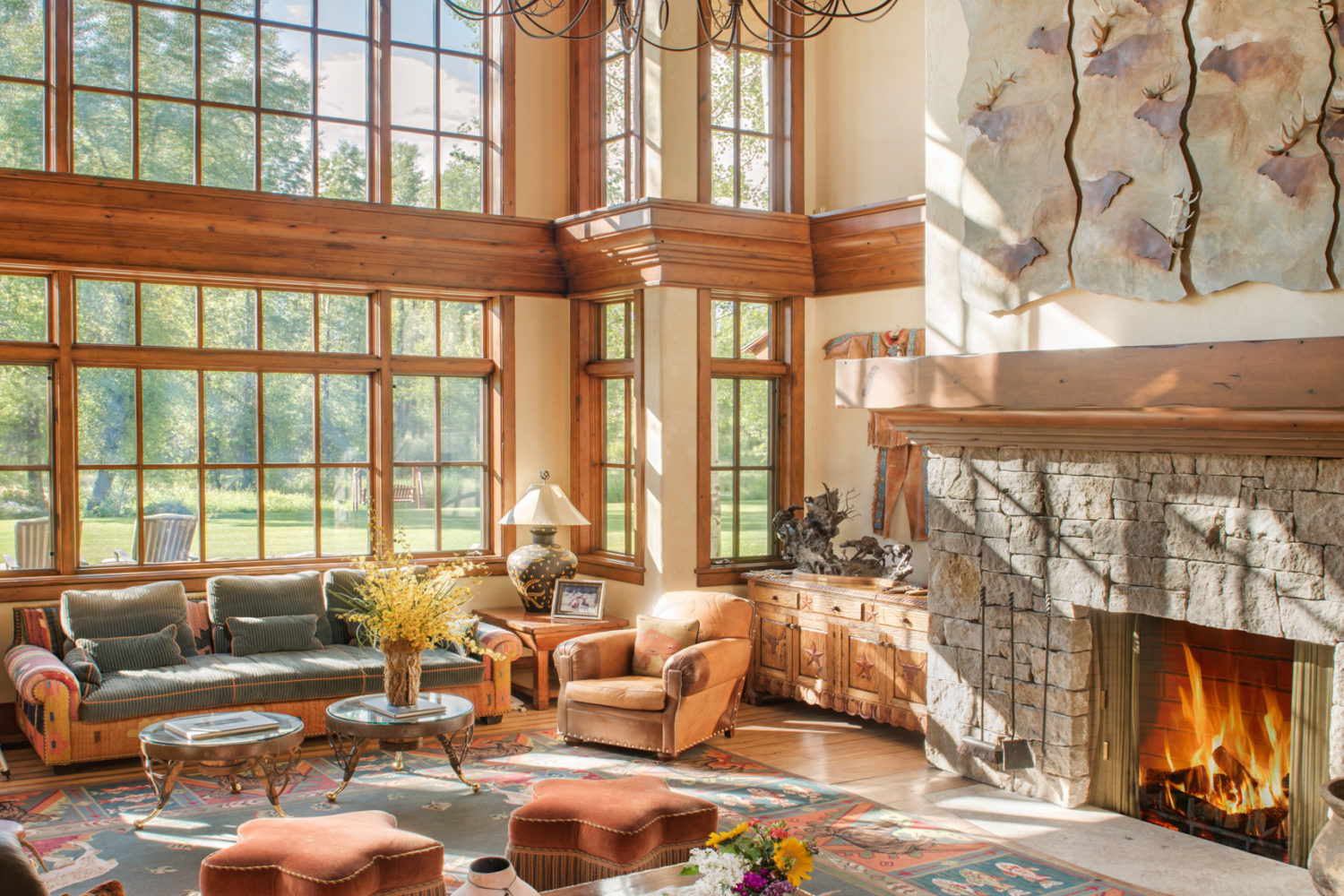 main_house_great_room A large sitting room with floor to ceiling windows, western art and furniture, and a lit fireplace