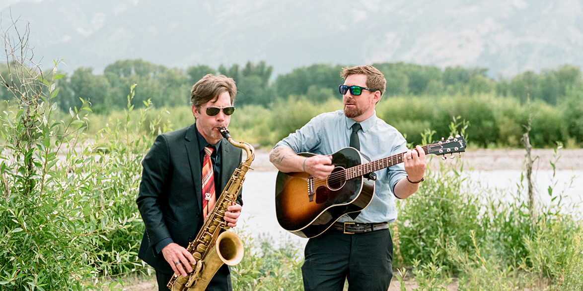 musicians_at_river A saxophonist and guitarist play music near a river in front of mountains